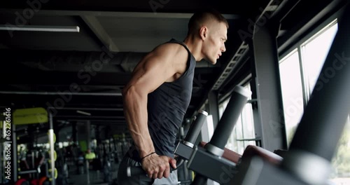 Young muscular man has workout at gym and pull up on training apparatus. Side view on athletic male doing dip exercise and training biceps muscles at fitness equipment in sport club