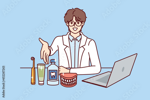 Man dentist pulls hand to screen, sitting at table with laptop and supplies for dental care. Young doctor dentist recommends purchasing toothbrush and toothpaste to maintain hygiene