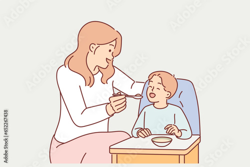 Mother feeds boy sitting in child seat with spoon, caring for kid and wanting to give son best