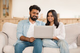 Happy young indian spouses relaxing with laptop on sofa, couple using computer, browsing internet or shopping online