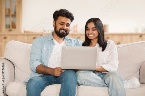Happy young indian spouses relaxing with laptop on sofa, couple using computer, browsing internet or shopping online