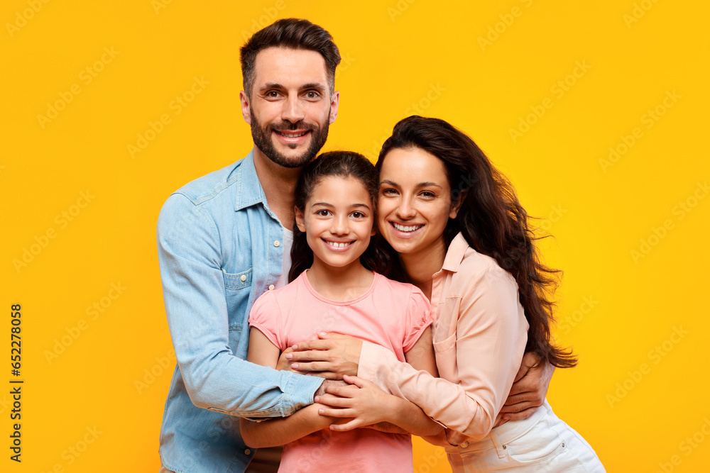 Happy european parents hugging their pretty daughter and smiling to camera on yellow background, studio shot - obrazy, fototapety, plakaty 