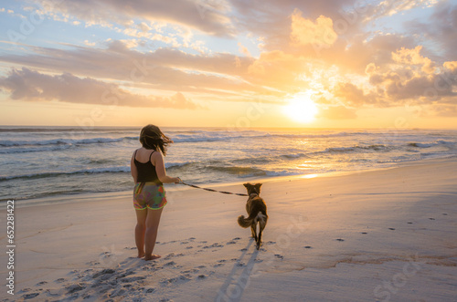 Female with her dog standing on a sand beach and enjoying the sunset