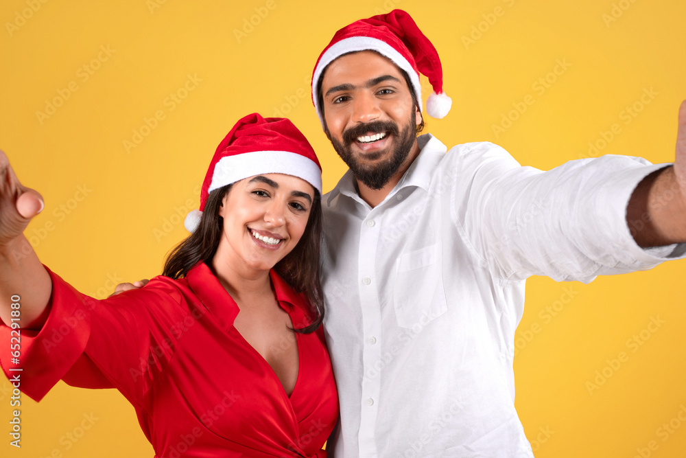 Smiling millennial arab man in Santa hat hug lady, taking selfie Smiling millennial arab man in Santa hat hug lady, taking selfie
