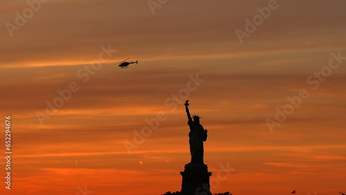 Tourists helicopter flying above Liberty Statue in Manhattan, against orange sunset sky. 4K travel video in New York.