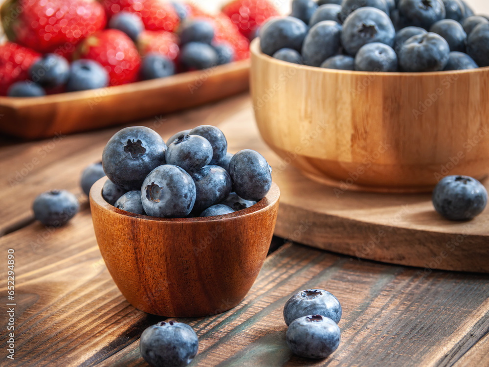 Blueberries close-up in a wooden cup on a dark brown wooden background in warm rays of light. Blueberries and strawberries in the background. Selective focus. Side view. Blueberries close-up in a wooden cup on a dark brown wooden background in warm rays of light. Blueberries and strawberries in the background. Selective focus. Side view.