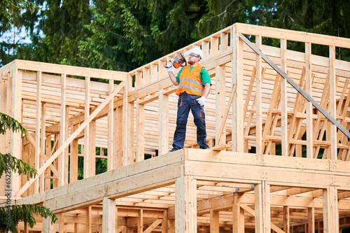 Carpenter building wooden frame two-story house. Bearded man wearing protective goggles carrying chainsaw on his shoulder, dressed in protective helmet, coveralls, and orange vest.