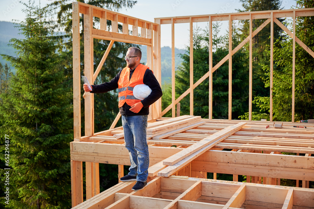 Manager overseeing construction of wooden two-story house located close to woods. Bearded man in glasses studying construction plan. Objective is to implement modern ecological construction methods. Manager overseeing construction of wooden two-story house located close to woods. Bearded man in glasses studying construction plan. Objective is to implement modern ecological construction methods.