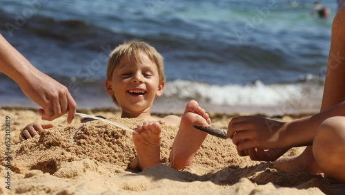 Child, tickling sibling on the beach on the feet with feather, kid cover in sand, smiling, laughing, enjoying some fun