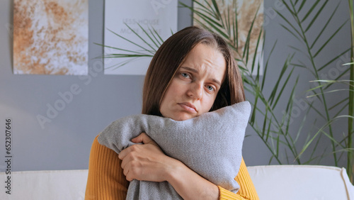 Sad unhappy worried bored woman sitting at home alone embracing pillow missing somebody being in bad mood looking at camera with upset facial expression.