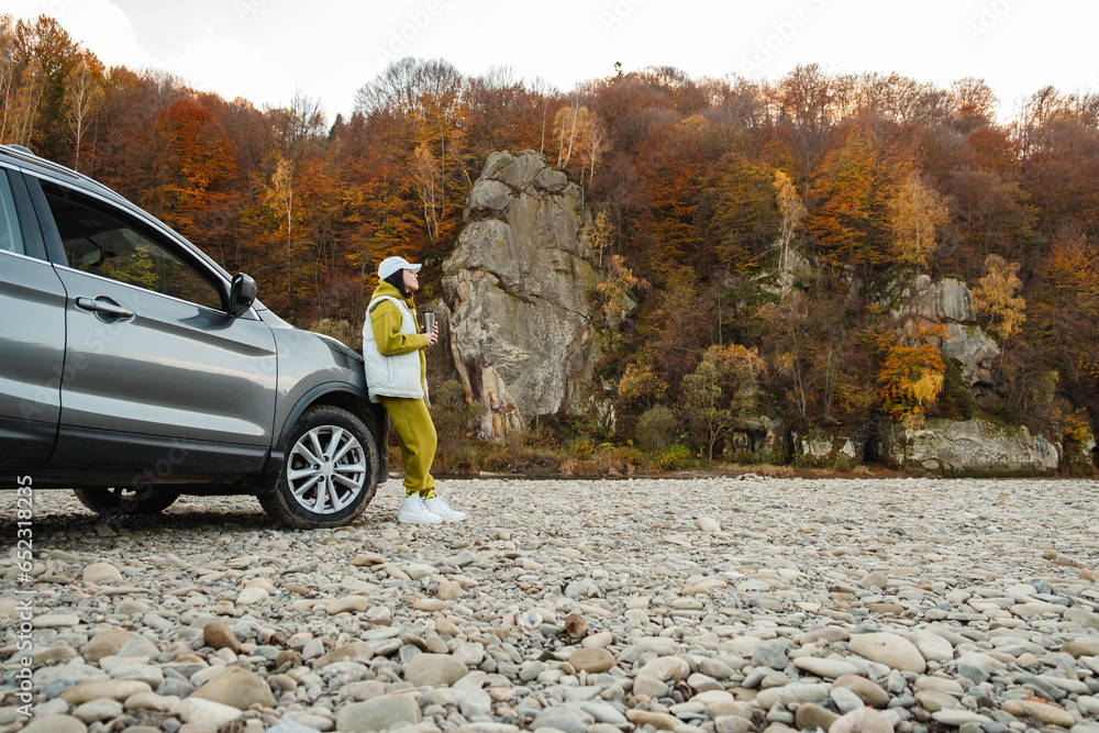 woman standing near car drinking tea looking at mountain river