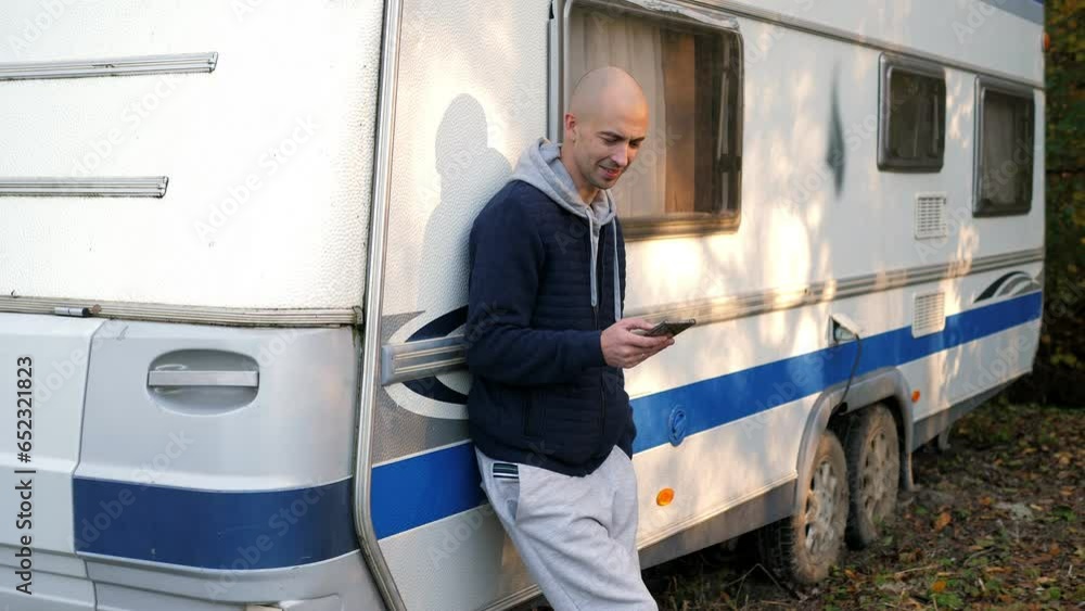 A smiling man with a phone in the autumn forest, he is leaning against the van. A man travels in autumn on a mobile home. A smiling man with a phone in the autumn forest, he is leaning against the van. A man travels in autumn on a mobile home.