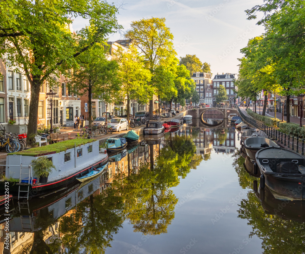 This photo showcases a peaceful Amsterdam canal scene. It includes an arched bridge, trees, boats docked in the canal and canal houses. Taken on a calm Sunday morning, the image reflects the canal's s