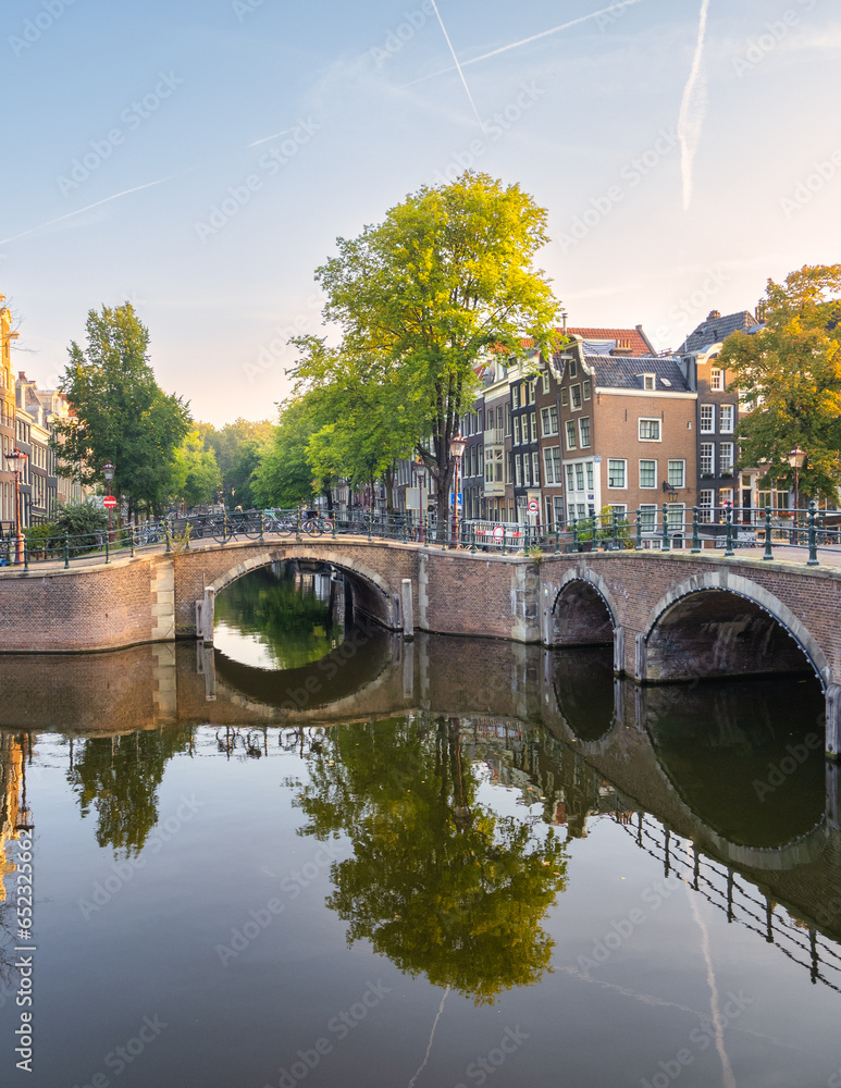 This photo showcases a peaceful Amsterdam canal scene. It includes an arched bridge, traditional canal houses, a few trees, and bicycles on the bridge railing. Taken on a calm Sunday morning, the imag