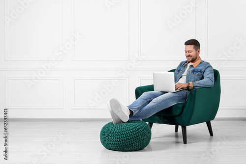 Happy man sitting in armchair and using laptop indoors, space for text
