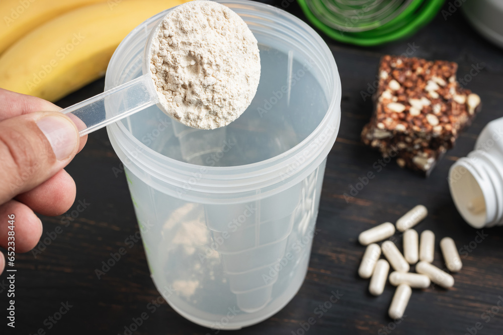 Male hand puts into a shaker scoop portion of whey, soy protein powder. White capsules of amino acids, vitamins, protein chocolate bar and bananas on a dark wooden board, bodybuilding food supplements Male hand puts into a shaker scoop portion of whey, soy protein powder. White capsules of amino acids, vitamins, protein chocolate bar and bananas on a dark wooden board, bodybuilding food supplements