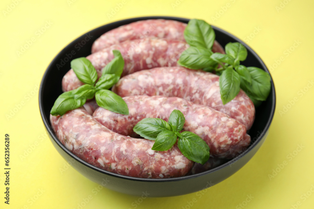 Raw homemade sausages and basil leaves in bowl on yellow background