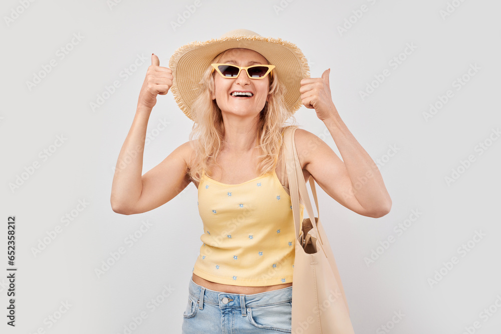 Portrait of a mature woman in summer clothes smiling joyfully showing thumbs up gesture isolated on white studio background. Approves good choice, right decision