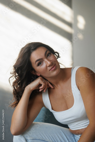 Beautiful brunette woman in white top and blue jeans sitting on blue armchair. Girl smile, happy. Portrait of young pretty woman. Smile and freshness. White background. Light from window, shadows
