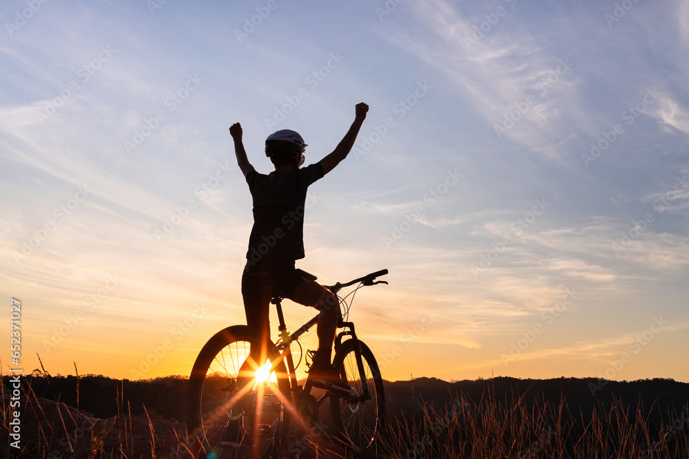 Obraz premium Silhouette of a cyclist on his mountain bike celebrating his victory after a challenging ride, with the sunset in the background