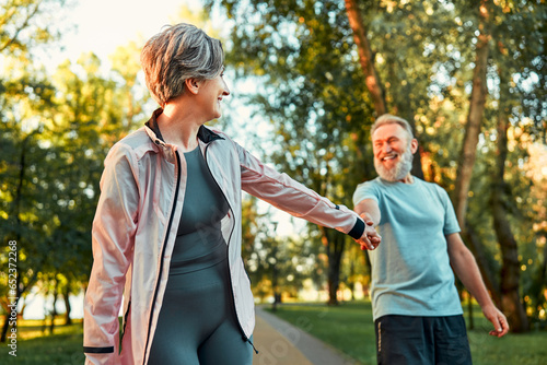 An older beautiful woman holds a man's hand and pulls him to sport training, and he stops and laughs. Lifestyle of the elderly. Activities in the fresh air.A healthy body is a healthy mind.