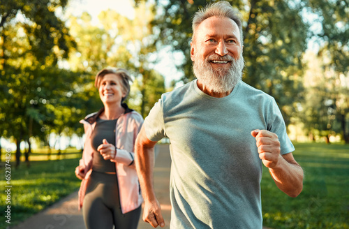 Fototapeta Naklejka Na Ścianę i Meble -  Cardio training. Close up of energetic fit sportsman spending morning in park and jogging together with wife. Active pensioners taking care of health and doing regular physical activities on nature.