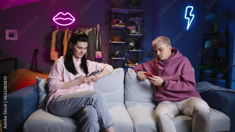 Fair-haired guy with dark-haired girl with phones on sofa in living room-studio. Expression of emotions. Girl is having fun, guy is unhappy. He lost in game, she won. Neon lights on backgrand