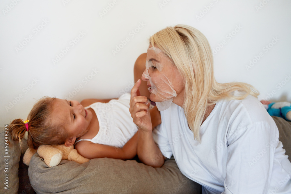 A young mother with a fabric mask on her face communicates with her daughter on the sofa at home. A young mother with a fabric mask on her face communicates with her daughter on the sofa at home.