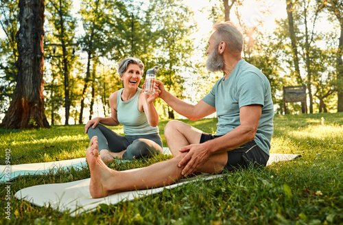 Sport and recreation outdoors. Angle view of two family partners having rest and sitting on fitness mats in park. Smiling senior lady sharing bottle of cold fresh water with husband.