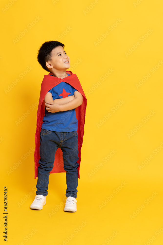 Superhero Asian boy standing and smiling with red cape and look aside isolated yellow background, Full body composition