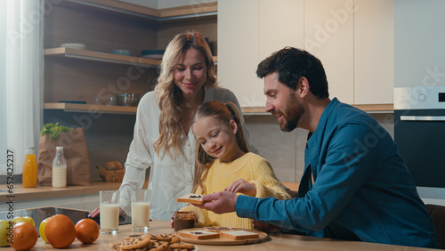 Happy Caucasian family preparing breakfast together talking at home kitchen mother hold glasses with milk little daughter child girl prepare toasts bread with chocolate pasta with father food delivery