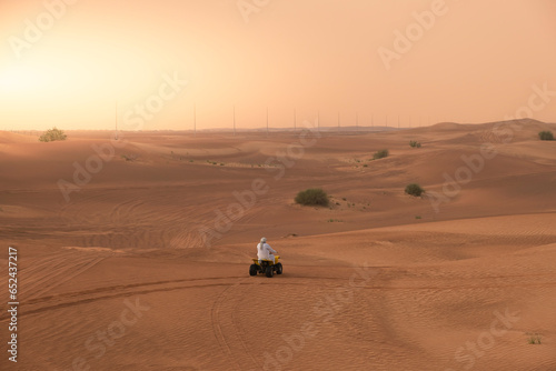 Arab man in traditional white robe outfit and kaffiyeh riding an ATV motorbike alone over sand dunes in the middle of the desert.