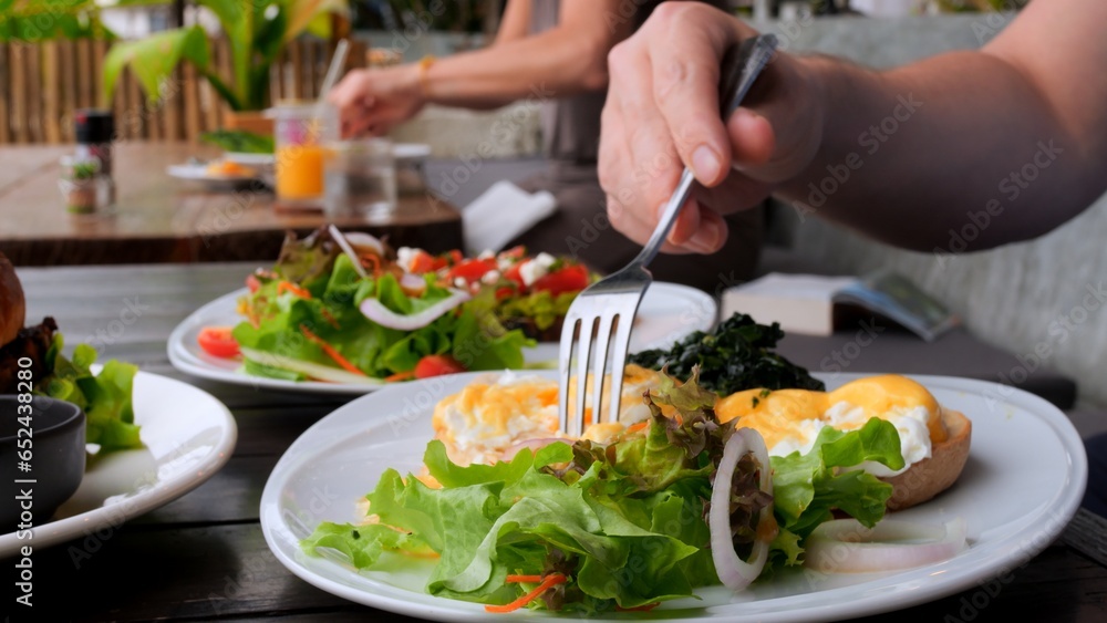 Man eating full meal as poached egg, salad and avocado toast in cafe. Close-up of man with fork pierces poached egg and eat tasty yolk. Order poached egg to eat healthy full meal. Full meal concept. Man eating full meal as poached egg, salad and avocado toast in cafe. Close-up of man with fork pierces poached egg and eat tasty yolk. Order poached egg to eat healthy full meal. Full meal concept.