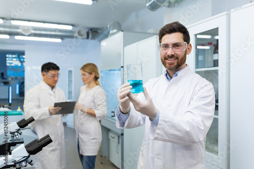 Portrait of a young male chemist standing in a laboratory, and holding a flask with an unknown blue substance, in his hands and smilingly looking at the camera.