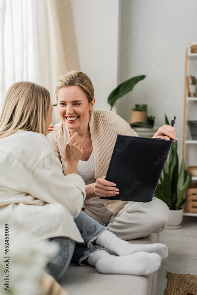 joyful attractive sisters in lovely outfits laughing and holding photo album, family bonding joyful attractive sisters in lovely outfits laughing and holding photo album, family bonding