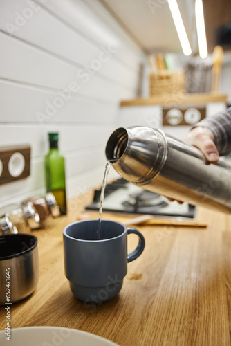 Vertical closeup of young man pouring hot coffee into cup from thermos while travelling with trailer van
