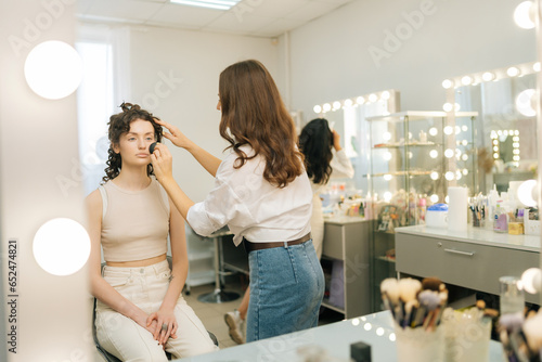 Reflection on mirror of unrecognizable female make-up artist applying cosmetic tonal foundation for face sculpting of pretty young woman in beauty studio. Concept of makeup and styling.