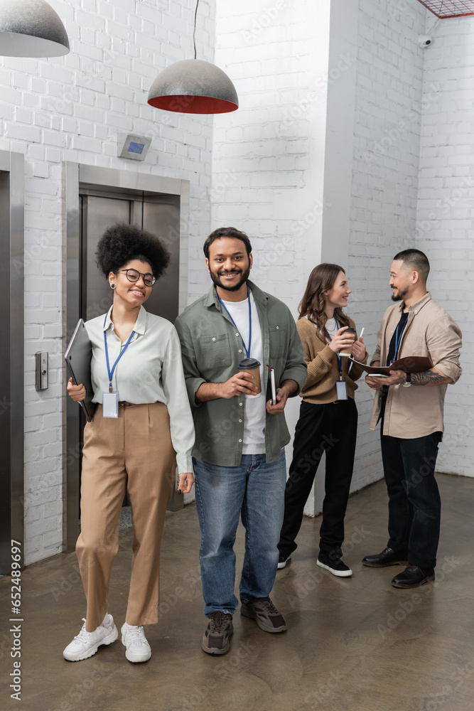 Fototapeta premium happy african american woman and indian man looking at camera near office elevators, diverse people