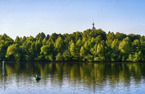 A fisherman on an inflatable rubber boat is fishing opposite an island overgrown with trees over which the dome of the Russian Orthodox Church rises