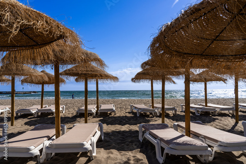 Tableau sur toile A sunny horizontal shot from under a row of thatched straw parasols on a sandy beach, looking out at empty white sun loungers, the blue sea, and a clear sky