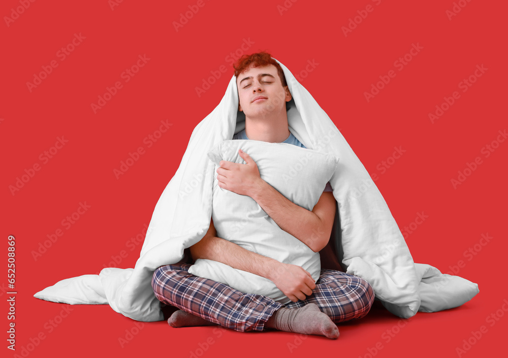 Young man with soft pillow and blanket sitting on red background Young man with soft pillow and blanket sitting on red background