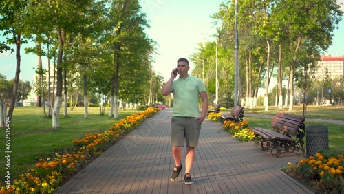 A young man walks in the park and talks on the phone. A man in a polo and shorts is walking along a green alley.