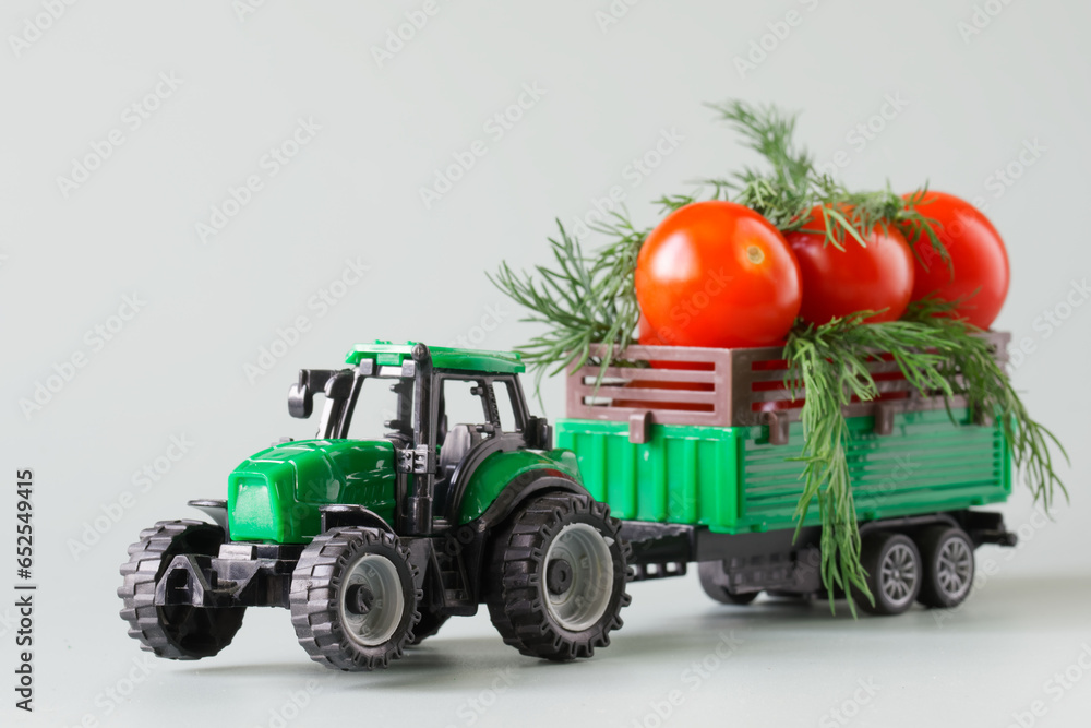 Obraz premium Green toy tractor carries fresh red tomatoes and herbs in a trailer. The concept of agricultural work, harvesting and delivery of crops. Toy world. Photo. Selective focus