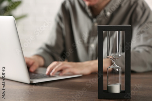 Hourglass with flowing sand on wooden table, selective focus. Man using laptop indoors