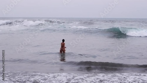 Woman in teeny bikini thong walks into grey Pacific Ocean waves