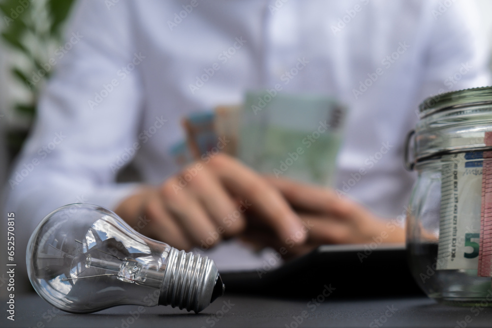 Fototapeta premium Electric light bulb. Man hands counting expenses bills on electricity banknotes of euro cash accounting on calculator. Close up of hands unrecognizable Businessman. High prices for energy. Save up