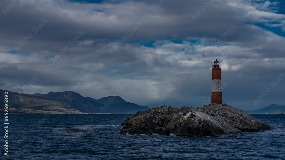 The famous southernmost old lighthouse Les Eclaireurs in the Beagle Channel. A stone tower with red and white stripes stands on a rocky island against the sky and clouds. Mountains in the distance The famous southernmost old lighthouse Les Eclaireurs in the Beagle Channel. A stone tower with red and white stripes stands on a rocky island against the sky and clouds. Mountains in the distance