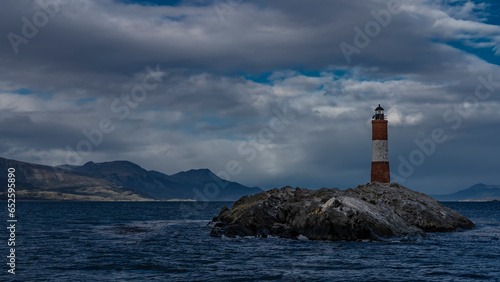 The famous southernmost old lighthouse Les Eclaireurs in the Beagle Channel. A stone tower with red and white stripes stands on a rocky island against the sky and clouds. Mountains in the distance