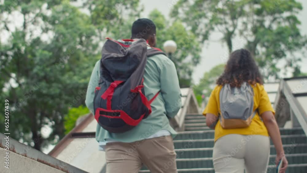 Medium rear follow shot of multiethnic man and woman with backpacks and tourist leaflets walking up stairs onto bridge or viewpoint, while exploring new foreign city during holiday Medium rear follow shot of multiethnic man and woman with backpacks and tourist leaflets walking up stairs onto bridge or viewpoint, while exploring new foreign city during holiday