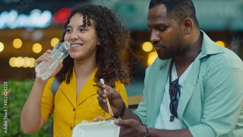 Medium close-up shot of African American husband and biracial wife eating delicious exotic street food while travelling abroad, exchanging impressions, unimpressed girl shaking head and drinking water photo